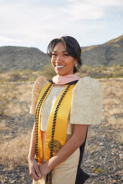 A portrait of Ashley Lazaro, who is standing in a field with gray rocks, brown grass and low, treeless hills in the background.. She is smiling with her hands clasped in front of her, and she is wearing a yellow dress embroidered with flowers that has short, puffy sleeves. Her dark hair that is pulled back with parted bangs and she has small silver hoop earrings. She is wearing an MPH stole, a yellow graduation stole and honors cord.