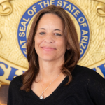 A photo of Beya Thayer, who is smiling. She has long brown hair and a black shirt standing in front of the seal of Arizona