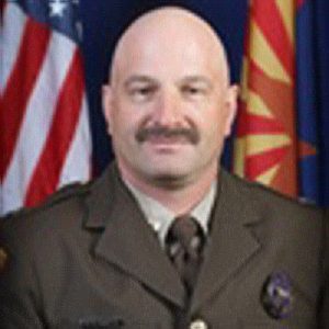 A photo of Captain Richard Martin smiling. He has no hair and a thick mustache with a brown county sheriff's uniform seated in front of the US flag and the Arizona flag in the background