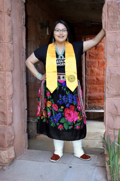 A portrait of DeeDee James who is standing in the exterior porch of a red stone building on campus. She has dark, straight hair pulled back, dark glasses, and is wearing a black v-neck shirt, a traditional Navajo black flowered skirt and moccasins. She is also wearing a yellow graduation stole, turquoise necklace, hanging earrings and bracelet. She is smiling and has her left arm up at head height and against a stone pillar of the building with irises in front. 