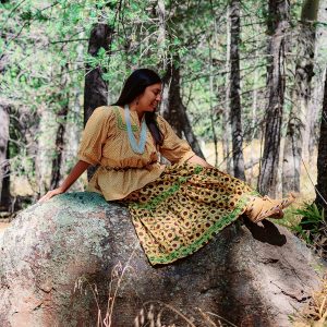 Marissa Tutt, wearing a traditional yellow dress and moccasins, posing on a boulder in a natural setting
