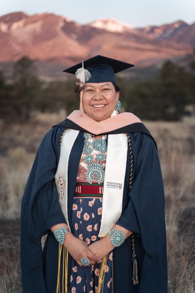 A portrait of Janet Yellowhair. She smiling and is standing in a field in Flagstaff at sunset with pine trees and the San Francisco mountains in the background. She is wearing a graduation cap and gown with a white and brown-tipped feather attached to the tassle. She is wearing two, long turquoise necklaces, large circle turquoise earrings, a large, round turquoise ring on her right hand and two large, round turquoise bracelets.