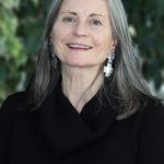 A portrait of professor Nicolette Teufel-Shone. She is smiling and has shoulder-length gray hair and is wearing a black turtleneck and large silver cross earrings.