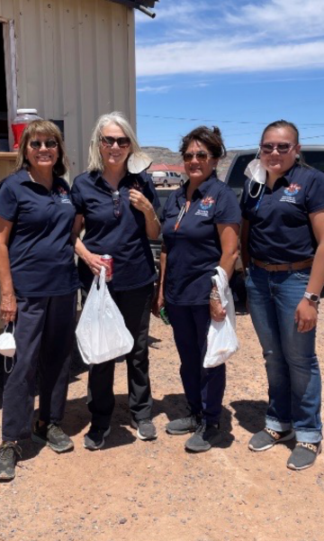 Four women wearing blue, short-sleeve, collared shirts with a Navajo Healthy Stomach Project logo pose outside for a photo on a sunny day. They are all wearing sunglasses.