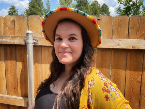 Photo of Ricky Camplain smiling and has long, dark hair. She is outdoors in front of a fence and is wearing a straw hat with a multicolored band around the rim, and a black shirt and a yellow flowered jacket.