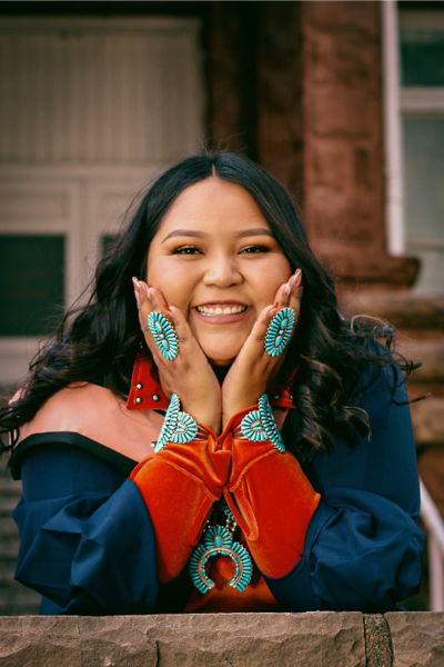 Portrait of Shawndeena George, who has long, dark hair that is parted in the middle and curled. She is wearing a graduation gown and a large turquoise necklace, a turquoise bracelet on each wrist and a large turquoise ring on each of her ring fingers. She is smiling and has her hands on her cheeks and her elbows propped up on a stone wall.. 