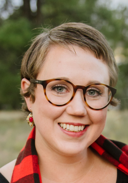 Photo of Shaylynn Shuler. She has short, blond hair parted on the side and is wearing round glasses, red and gold dangling earrings and a red plaid button-down shirt. She is smiling.