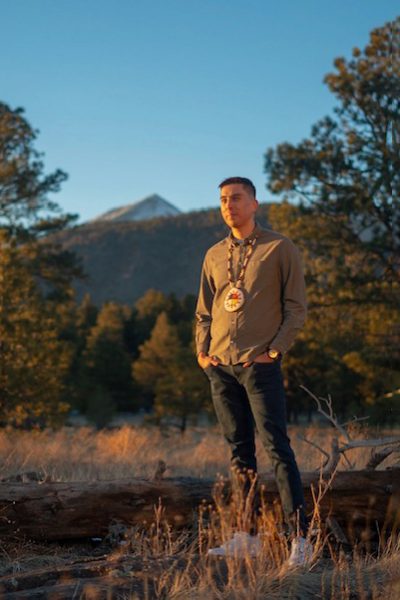 A portrait of Skyler Bordeaux standing in a field in Flagstaff with the San Francisco Peaks in the background. He is wearing a long, beaded necklace with his tribe's symbol and a light brown button-down shirt, black jeans and white running shoes. He has short, dark hair.