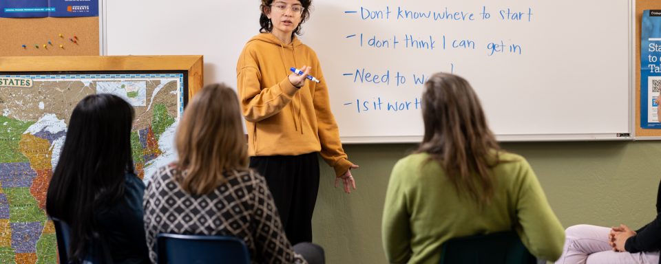 A CUPI Participant in a classroom outlining obstacles to higher education on a whiteboard, facing a panel of three women in front of them.