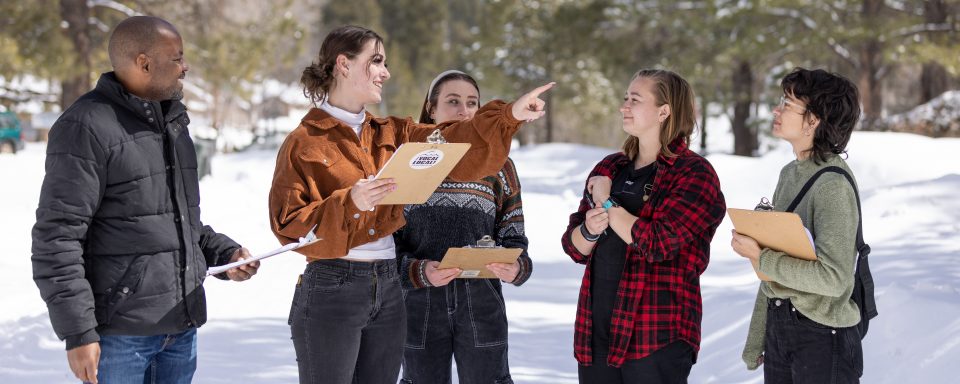 A group of CUPI students out in a snow covered forest, holding clipboards and smiling