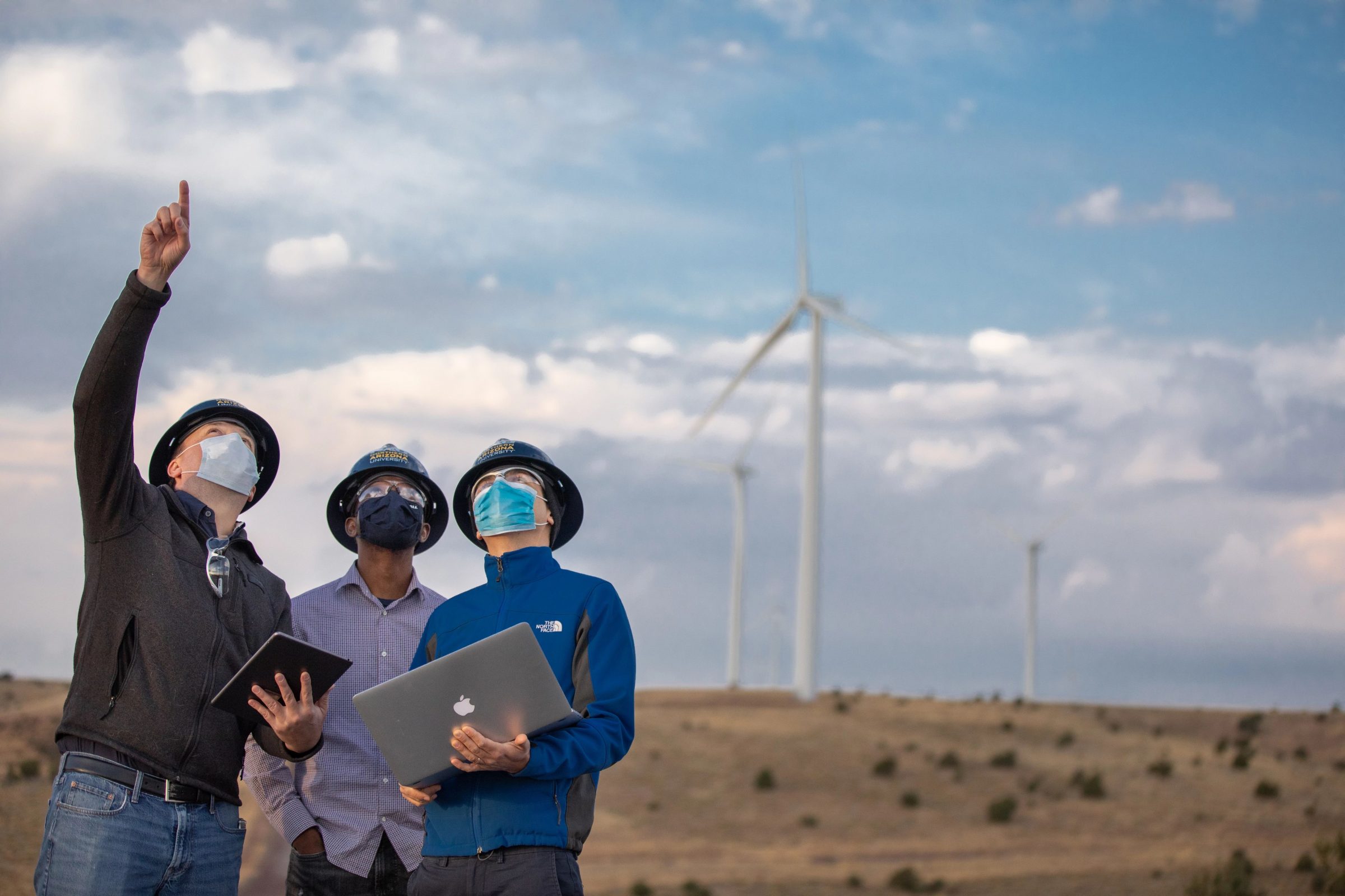 Three people looking up at a wind turbine--one person is pointing up with their arm extended-- behind them, in the distance, are other turbines.
