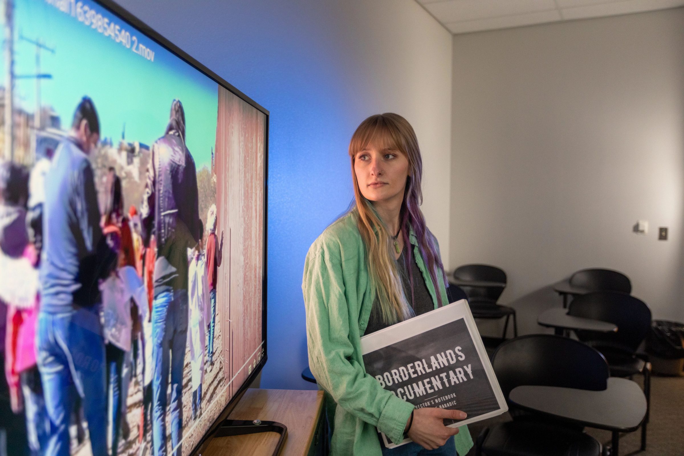 Mackenzie Baradic standing beside a large television screen while presenting her documentary film.