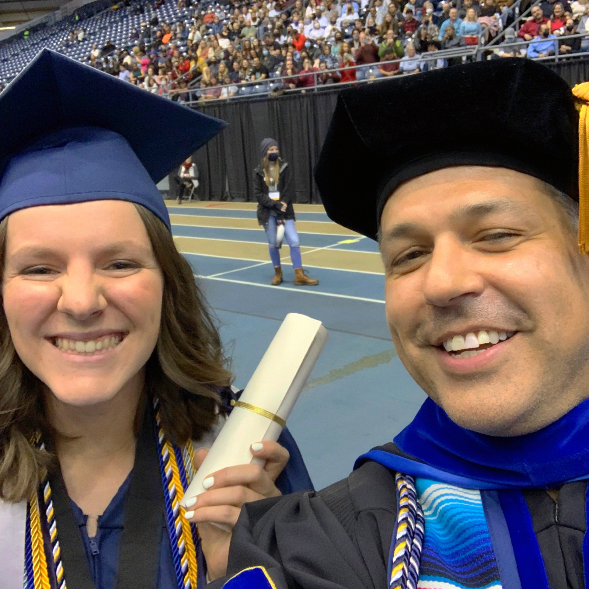 Selfie of Lizy Martinez, wearing cap and gown while holding a diploma at commencement, beside Professor T Mark Montoya.