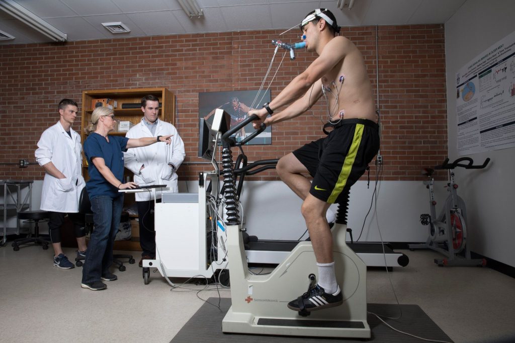 Students and Professor Traustadóttir look at results on computer screen as study subject rides stationary bicycle while hooked up to monitors.