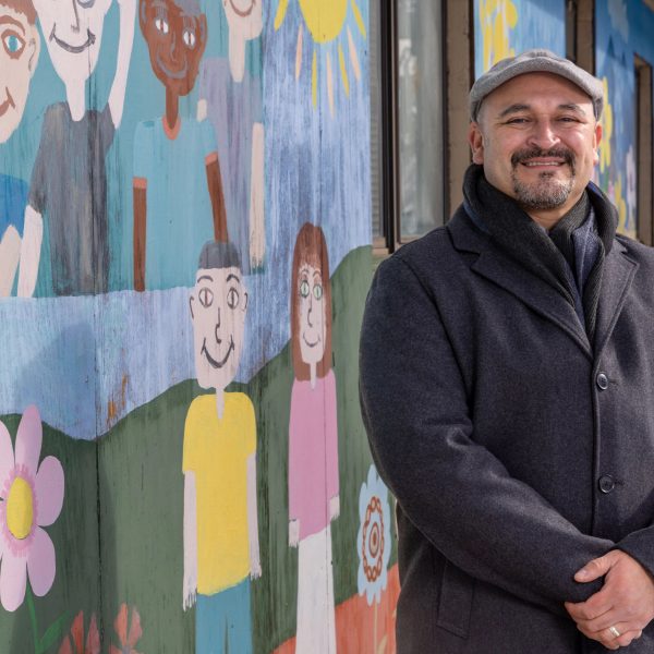 NAU alumnus and Coconino County Board of Supervisors vice chair Jeronimo Vasquez stands beside mural of smiling children, flowers, butterflies, and the sun. Vasquez is looking at the camera and smiling.