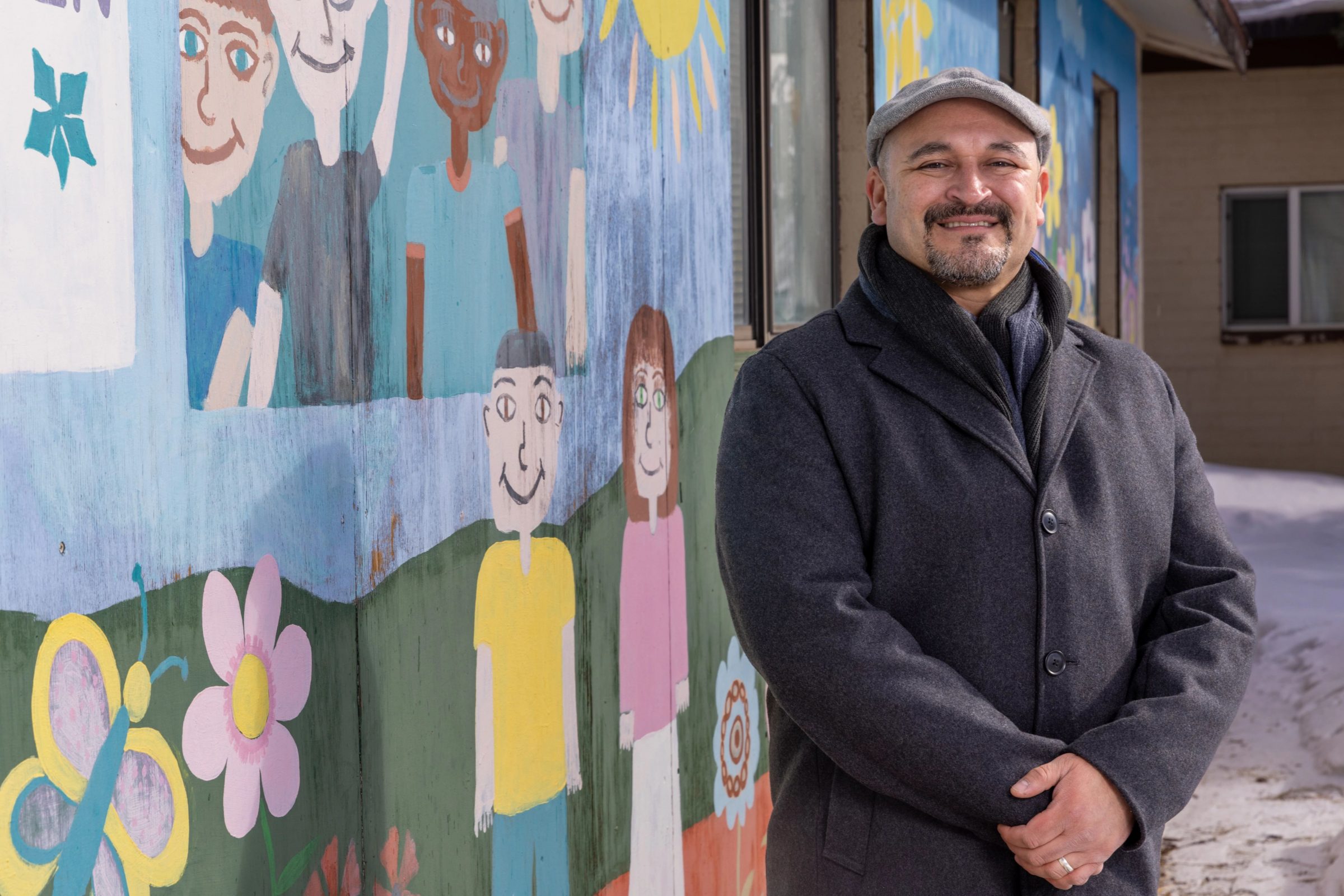 NAU alumnus and Coconino County Board of Supervisors vice chair Jeronimo Vasquez stands beside mural of smiling children, flowers, butterflies, and the sun. Vasquez is looking at the camera and smiling.