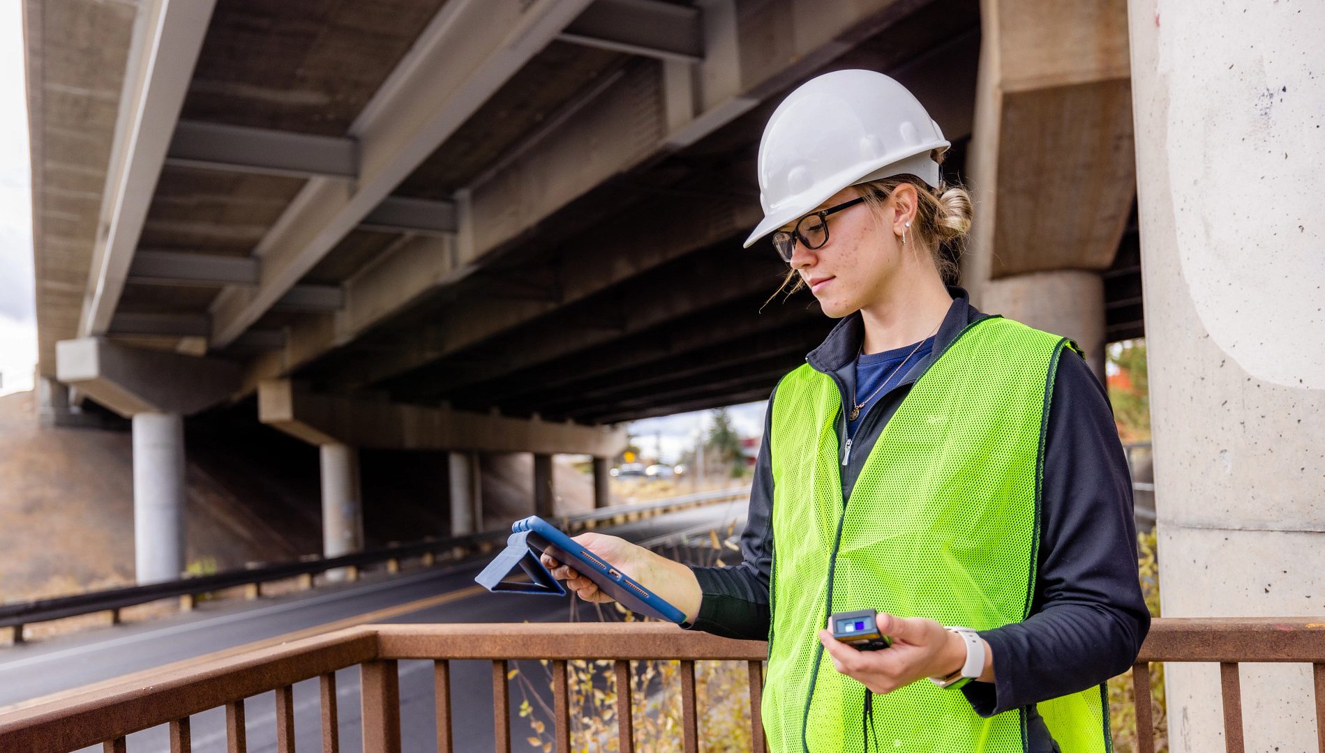 Civil Engineer major Jenna Hays, wearing a safety vest and hard hat, stands under bridge while looking at data on hand-held equipment.