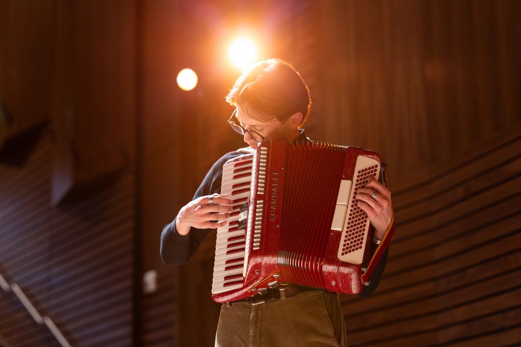 Honors Music Education and Music Performance major Will Whitten looking down while playing a red accordion, with studio light shining from behind.