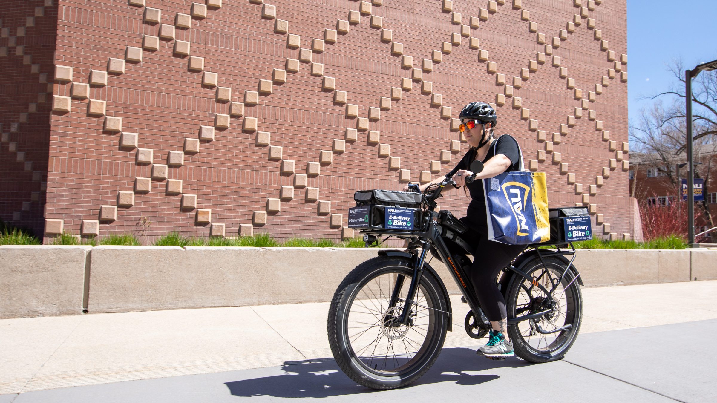 Employee rides the N A U ’s Printing Services’ delivery e-bike carrying a bag with the N A U logo. The bike has storage boxes above the front and back wheels labeled, “e-delivery bike.”