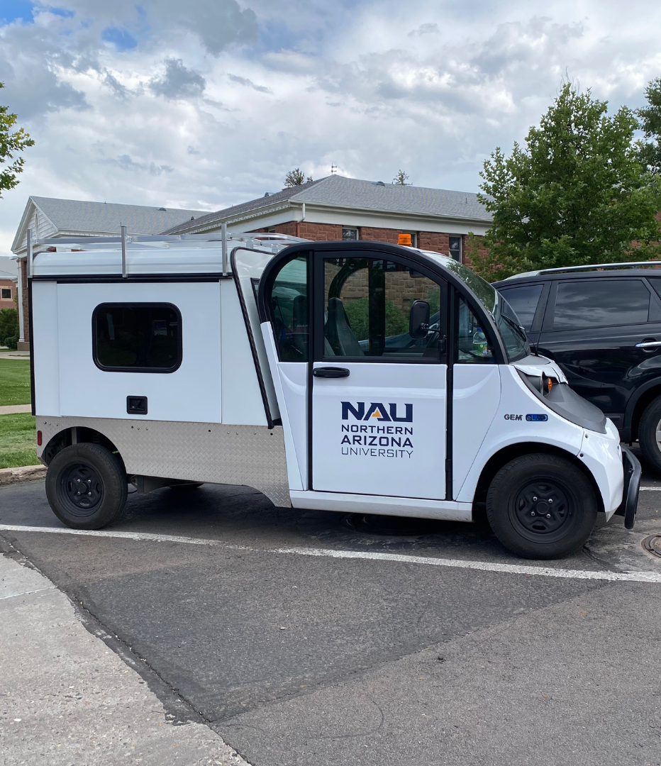 A parked white Electric Utility Vehicle (EUV) with the logo N A U Northern Arizona University on the door and a utility shell on the back for storing tools.