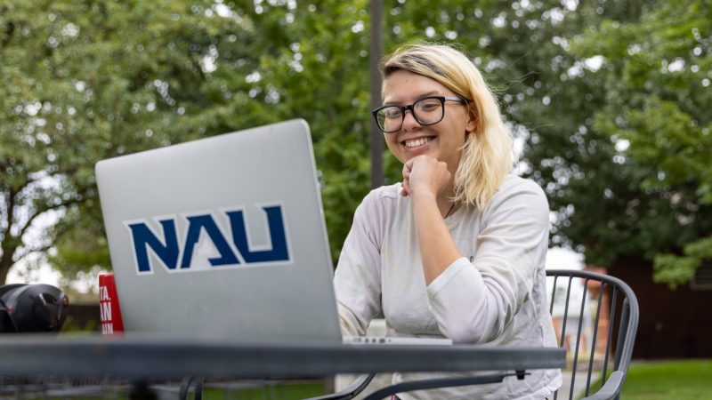 A student smiling as she works on a computer at a table outside.