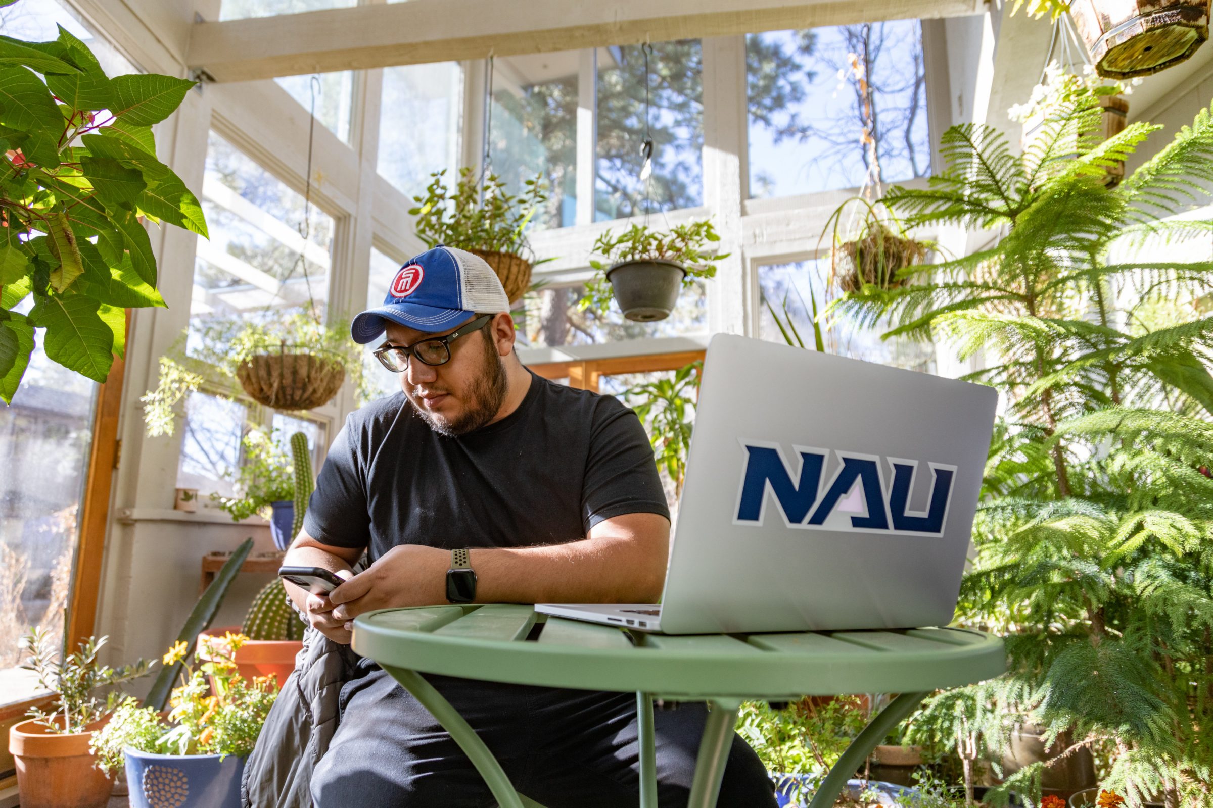 A student looks at his phone and laptop while sitting in a greenhouse surrounded by plants.
