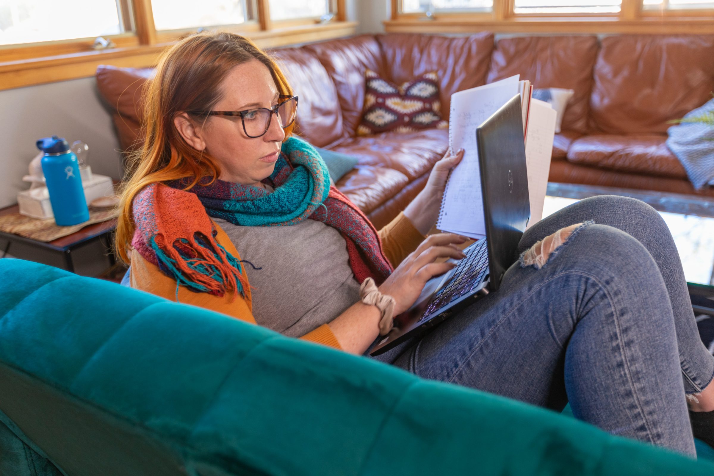 A student studies on the couch at home with a laptop and notebook.
