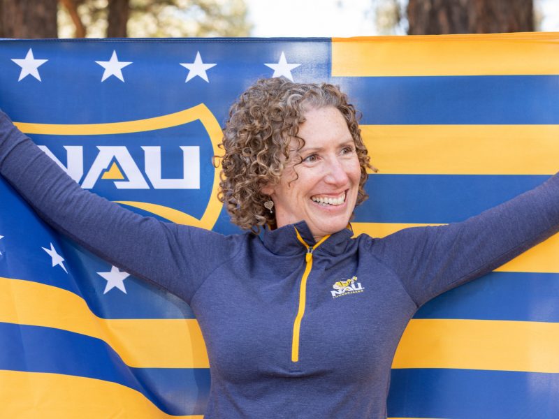 A person smiles while proudly holding an N A U flag and wearing N A U gear.