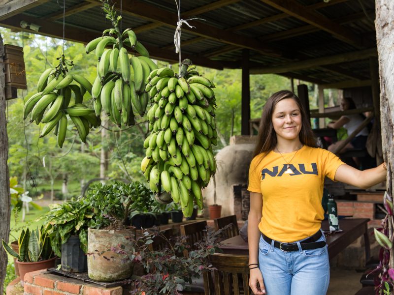 Student wearing orange Northern Arizona University t-shirt standing beside bananas while studying abroad.