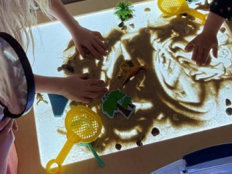 Two children standing at a light table with sand poured over it. Boy is holding a magnifying glass while the girl spreads her hands on the table.