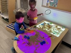 Two children playing at a light table with sand poured over it. One light is purple and the other is a white light. Children are holding a sifter and a microscope.