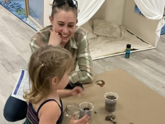 A mom watching her child show her a sample of rocks in a jar, next to other samples of dirt and sand.