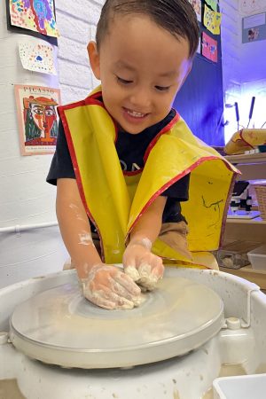 Child smiling using his hands to mold clay on a pottery wheel.