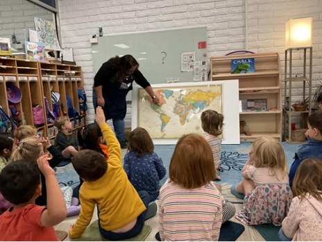 A teacher showing a world map, with children sitting on carpet squares watching.