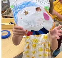 A child holding a paper mask with eyes cut out, decorated with crayons and five small shapes.