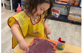 A child wearing a smock, combining paint on construction paper to make a large purple circle.