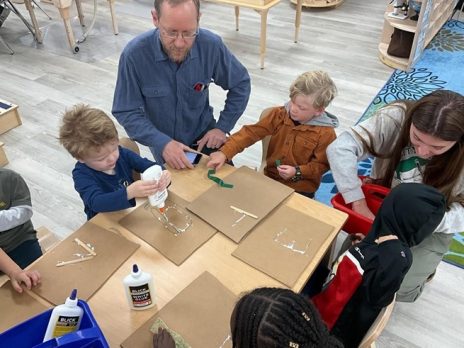 A teacher with students at a table, gluing popsicle sticks onto cardboard to start a collage.