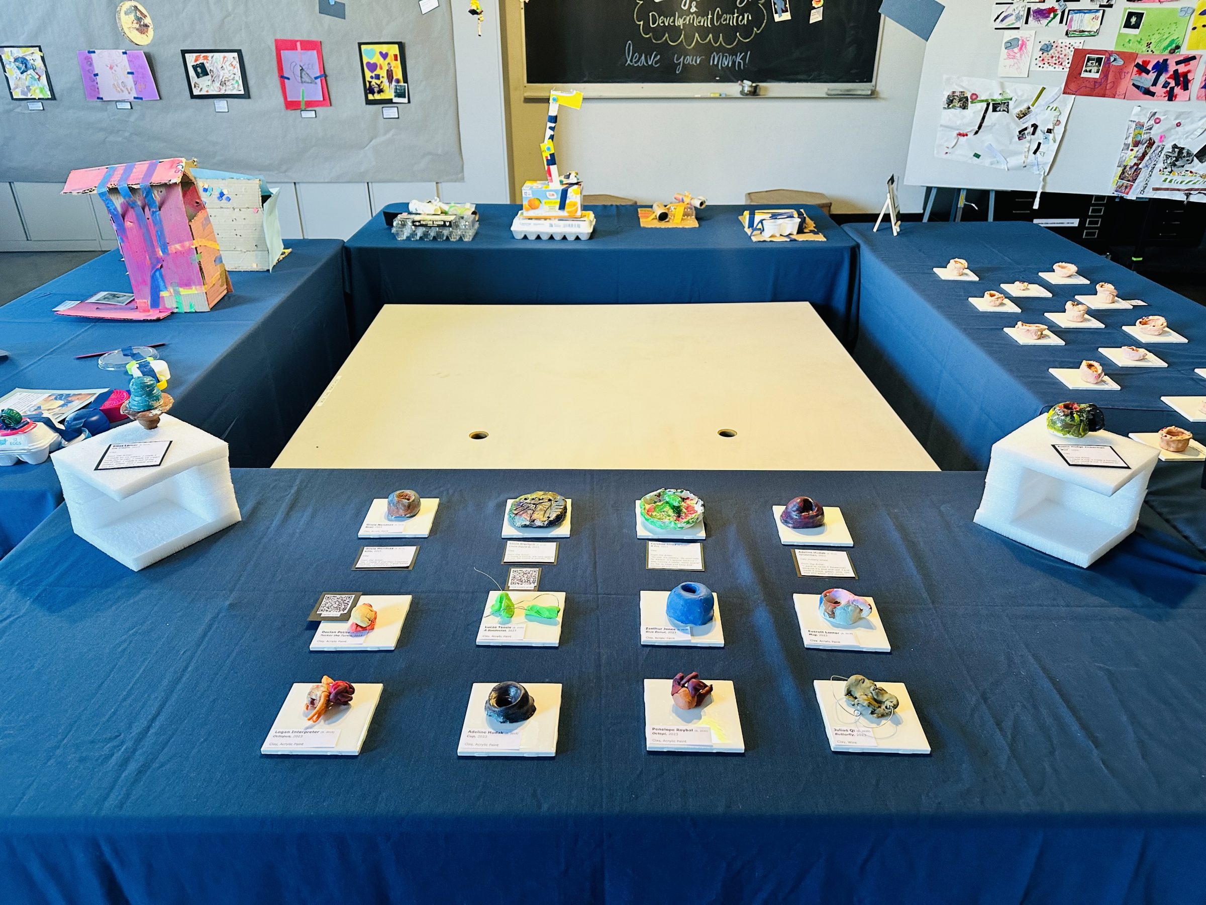 Clay puts made by children displayed on individual white tiles on a blue table.