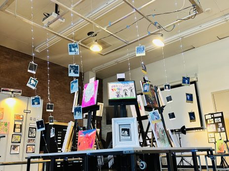 Display of children's art work in a gallery, with framed pictures and hanging strings of photos.