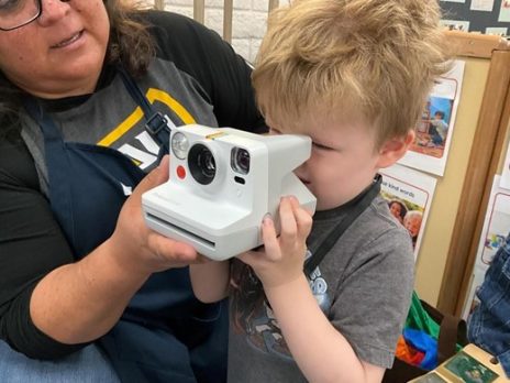 A teacher helping a child hold and point a poloroid camera.