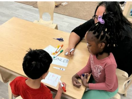 A teacher with two children writing with markers at a table.