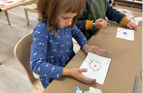 A child sitting at a table with an art piece she crteated. It has small dots in different colors, forming circles around the center.