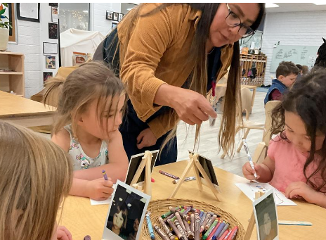 A teacher giving directions to children on making a self portrait with crayons. Each child has a polaroid displayed in front of them.