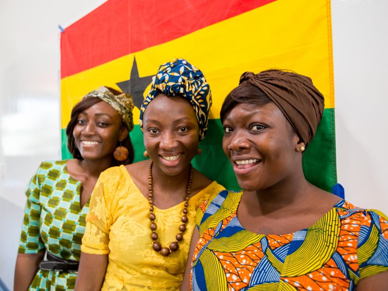 3 students smiling in front of the flag of Ghana.