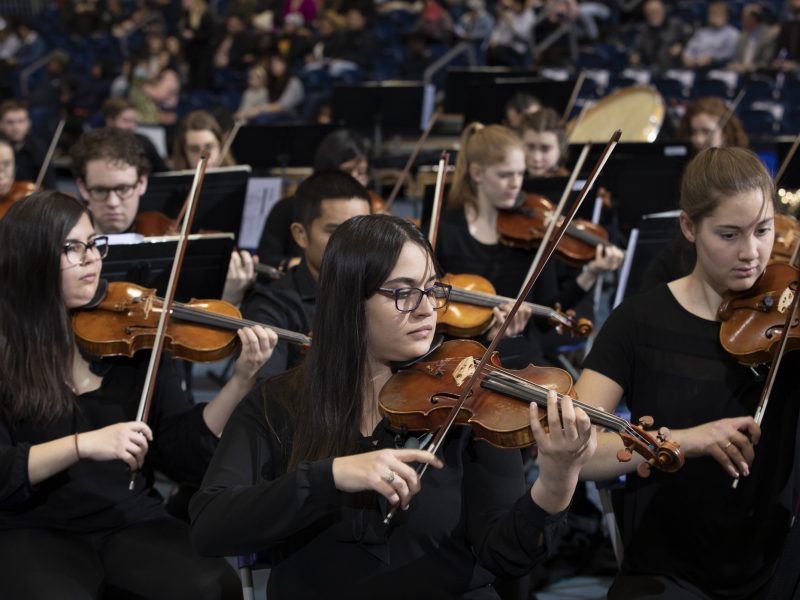 Students playing violins in an orchestra on stage.