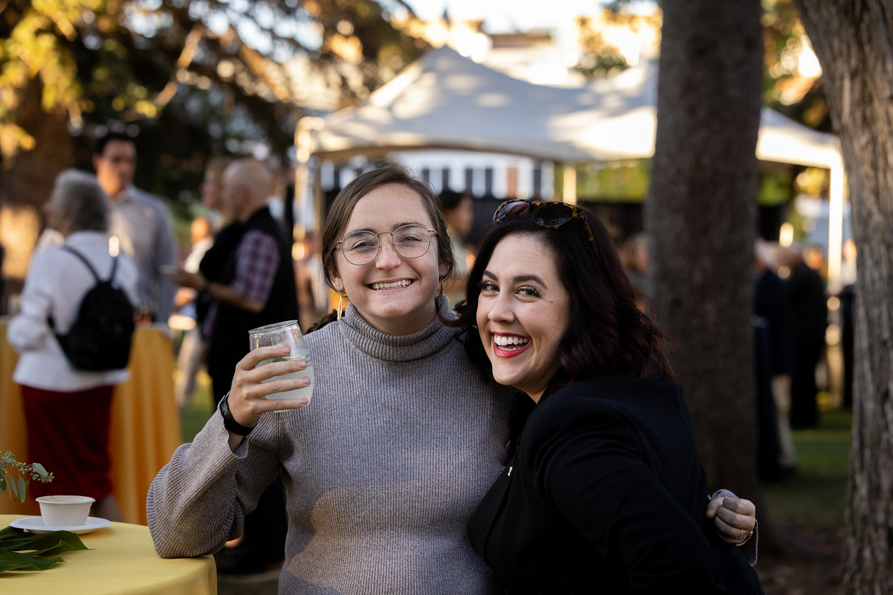 Two women pose for a selfie in front of a crowd of outdoor conference attendees at sunset in a grassy quad.