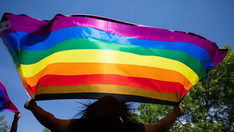 Pride flag being held up against a blue sky.