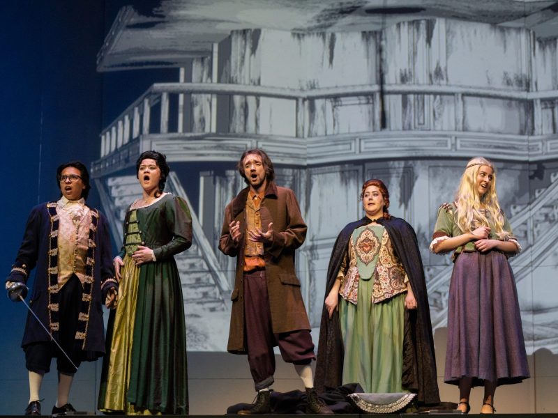 A group of opera performers dressed in character and singing on a stage.