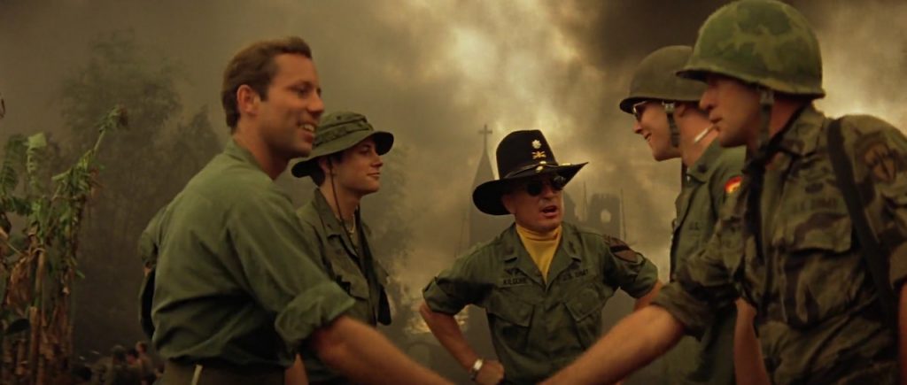 War scene from "Apocalypse Now" - soldiers in fatigues shaking hands in front of an officer in a calvary hat. Steeple of a church burns in background.