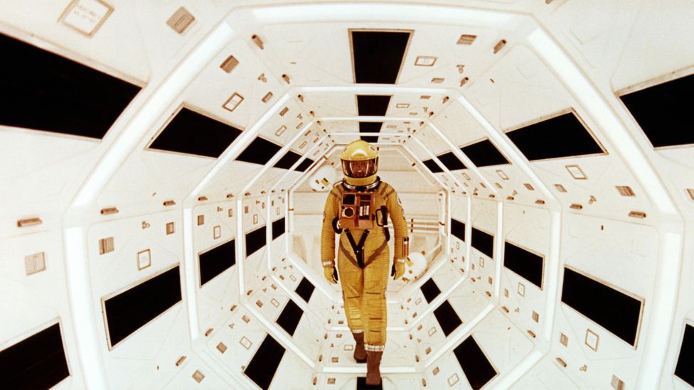 An astronaut in old school space suit stands in a tunnel connecting two sections of a stylish and symmetrical space ship.